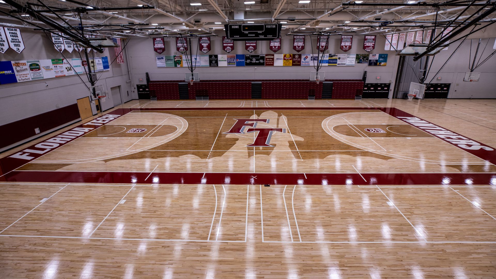 An overhead view of the basketball court at Florida Tech, featuring a polished wooden floor with the Florida Tech logo at center court, surrounded by red and white court lines, and bleachers in the background